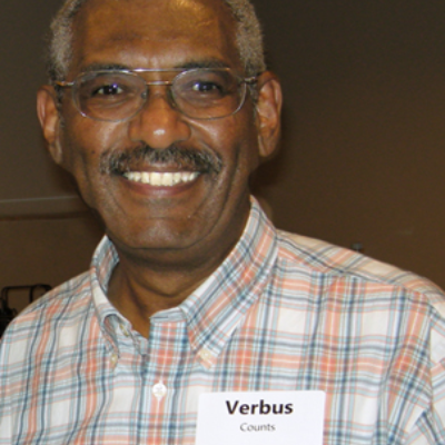 Smiling senior African Amercian Man with short grey hair and glasses wearing a checked white and peach shirt with a name badge reading Verbus Counts